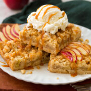 Three apple pie crumb bars stacked on a plate with a scoop of vanilla ice cream on top and drizzled with caramel sauce. - 1