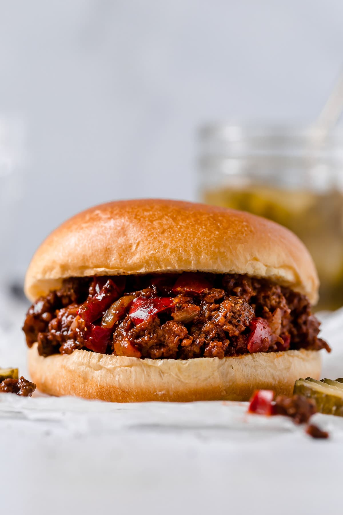 Close up image of a sloppy joe in a hamburger bun, it's sitting on a white tabletop and parchment paper with a jar of pickles in the background.