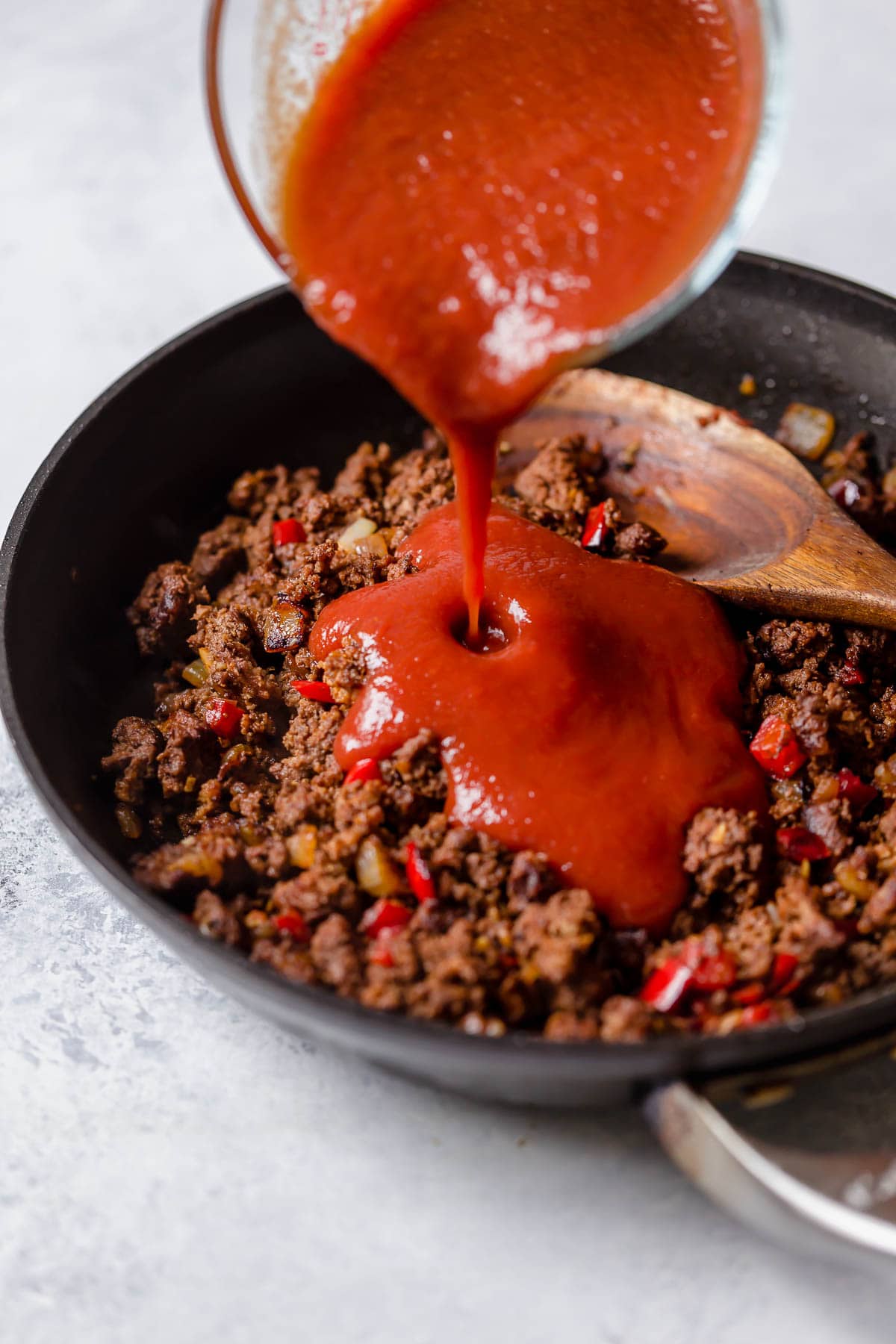 Pouring tomato sauce into ground beef, bell pepper and onion mixture in skillet. 