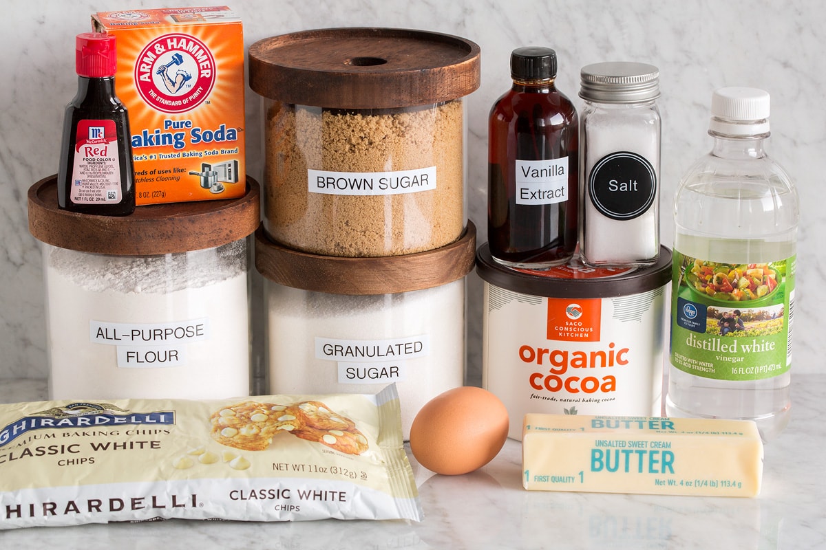 Image of ingredients used to make red velvet cookies. Includes flour, granulated sugar, brown sugar, cocoa powder, baking soda, red food coloring, vanilla extract, salt, vinegar, butter, egg and white chocolate chips.