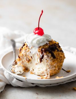 Fried ice cream cut into to show ice cream center. It is drizzled with chocolate sauce, topped with whipped cream and a maraschino cherry.