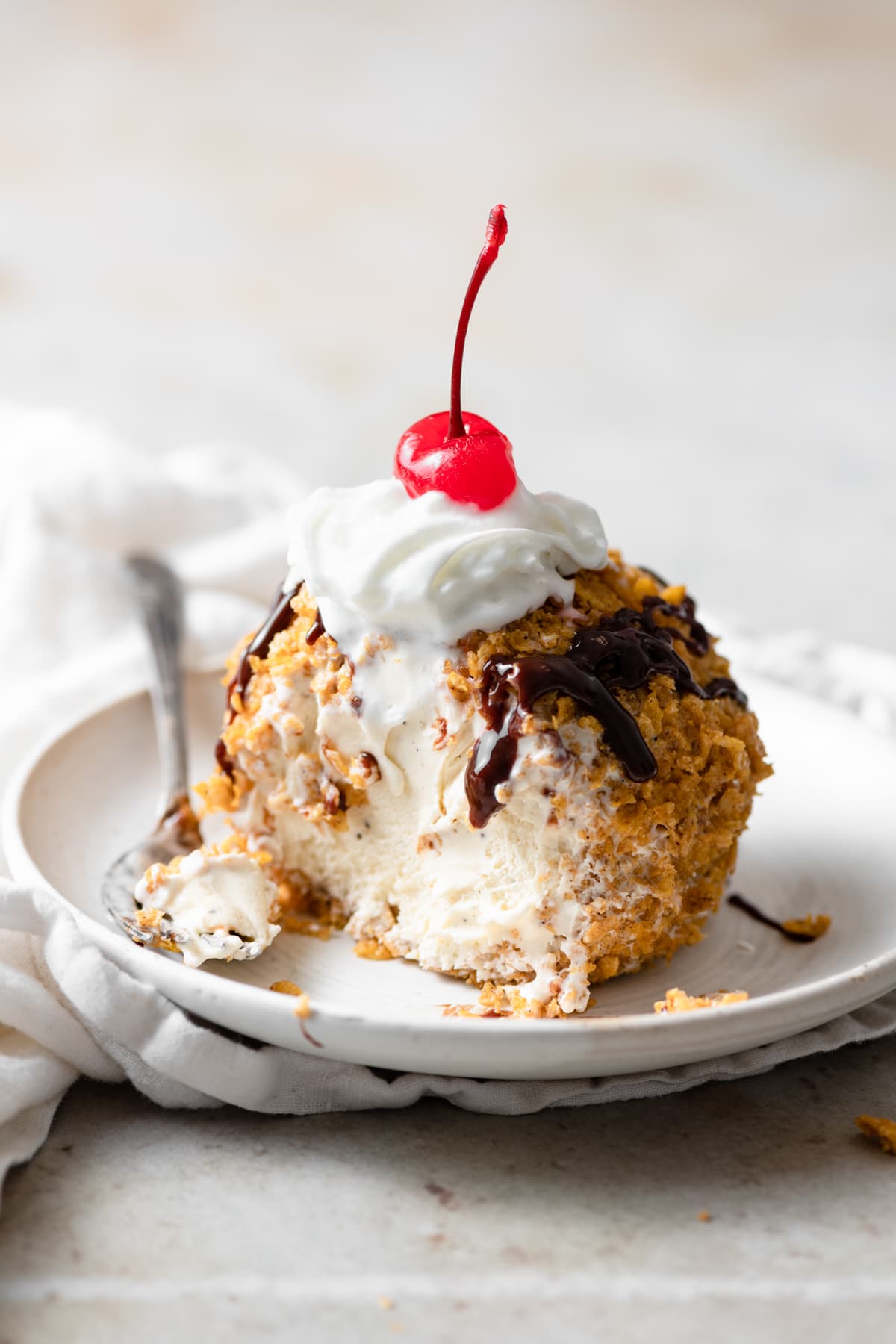 Fried ice cream cut into to show ice cream center. It is drizzled with chocolate sauce, topped with whipped cream and a maraschino cherry.