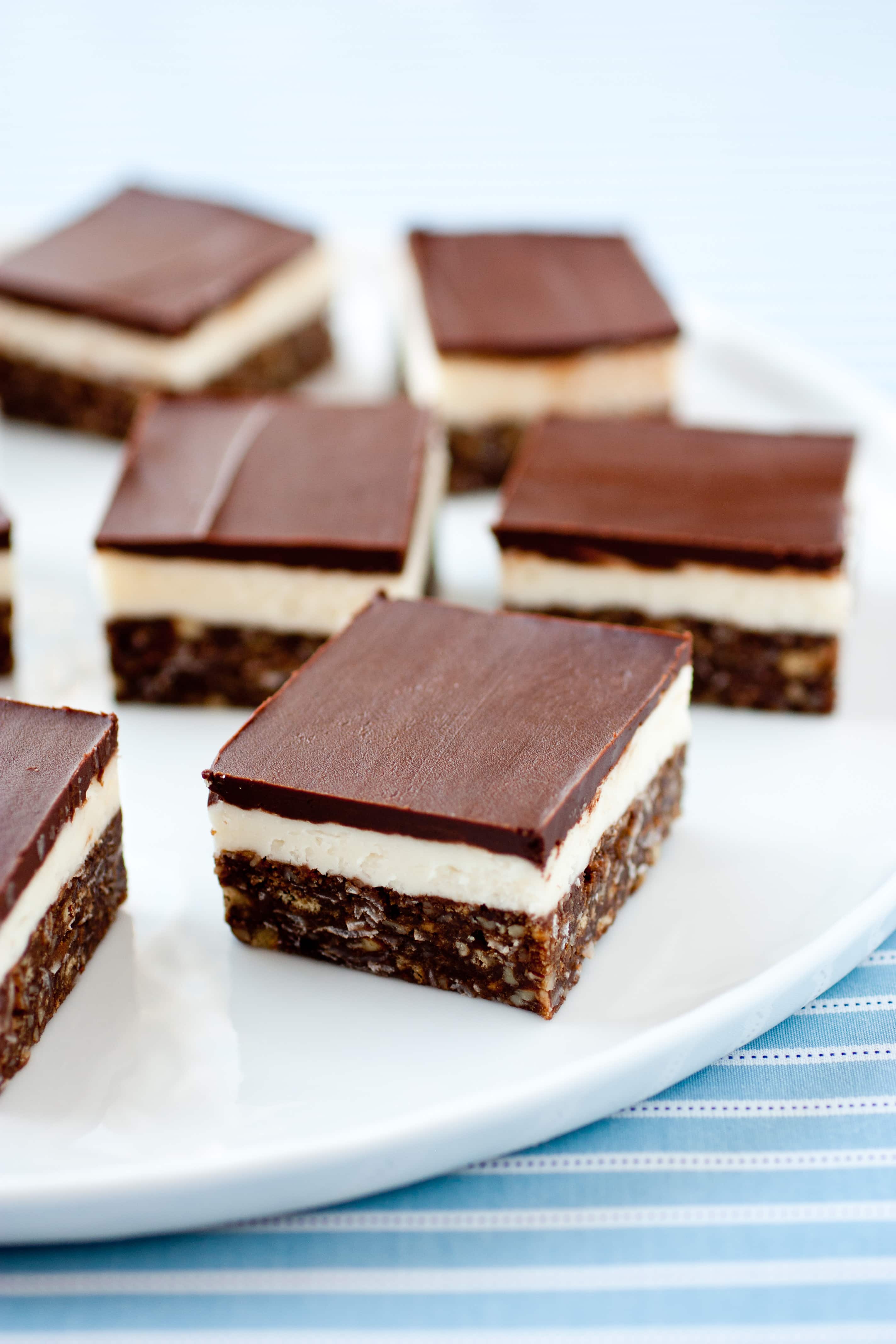 Nanaimo Bars shown on a white serving platter set over a blue striped cloth.