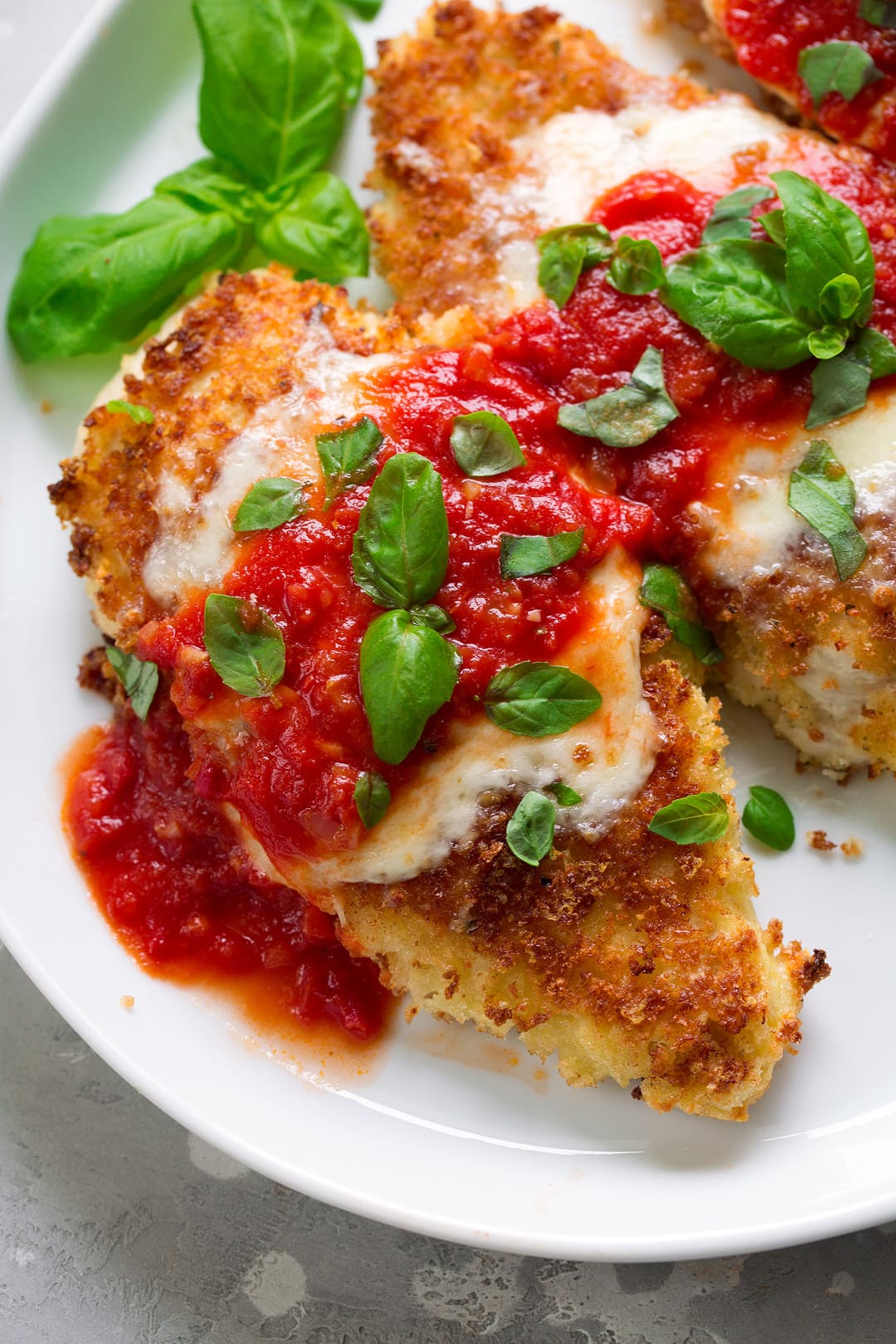 Overhead close up image of two pieces chicken parmesan on a white serving plate. Chicken is covered with golden brown Panko bread crumbs, melted mozzarella, marinara sauce and fresh basil.