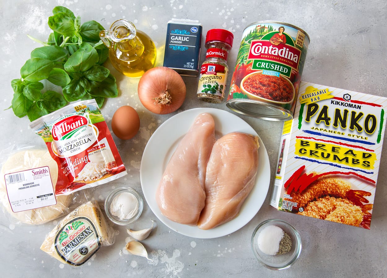 Ingredients needed for chicken parmesan shown here including panko, chicken breasts, crushed tomatoes, oregano, dried and fresh garlic, onion, egg, olive oil, basil, mozzarella, provolone, parmesan, and flour.