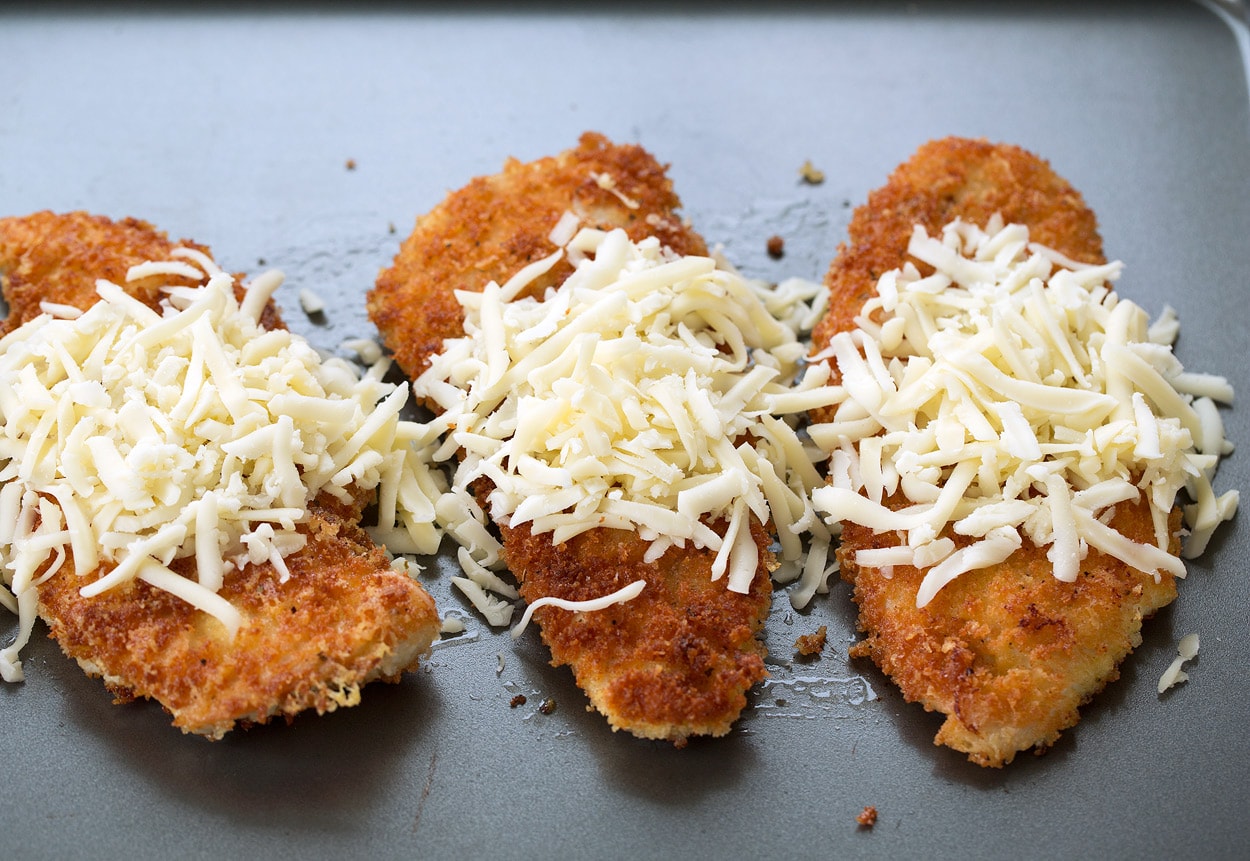 Placing browned chicken cutlets on baking sheet then covering with shredded cheeses.