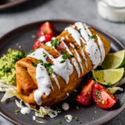 Chicken Chimichanga on a plate. It's drizzled with sour cream, garnished with cilantro and served with tomatoes and guacamole on the side.