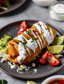 Chicken Chimichanga on a plate. It's drizzled with sour cream, garnished with cilantro and served with tomatoes and guacamole on the side.