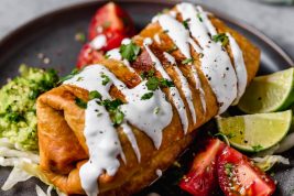 Chicken Chimichanga on a plate. It's drizzled with sour cream, garnished with cilantro and served with tomatoes and guacamole on the side.