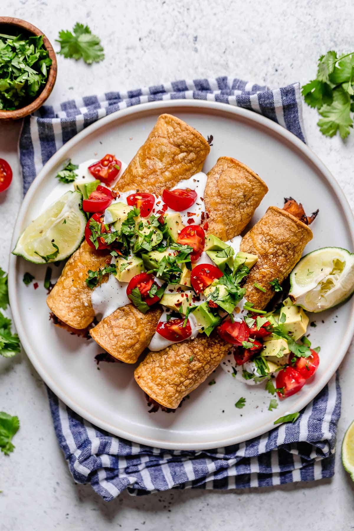 Taquitos on a white plate topped with sour cream, tomatoes, avocados and cilantro. Lime wedges are set of to the side and plate is set on a blue striped napkin.