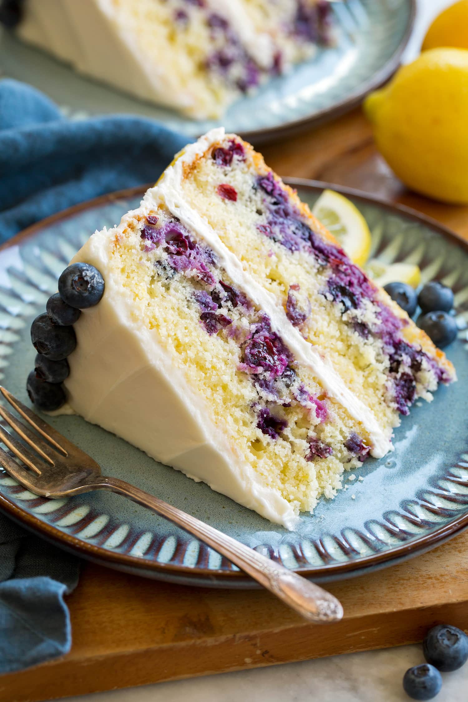 Slice of lemon blueberry cake with cream cheese frosting on a blue plate set over a wooden platter.