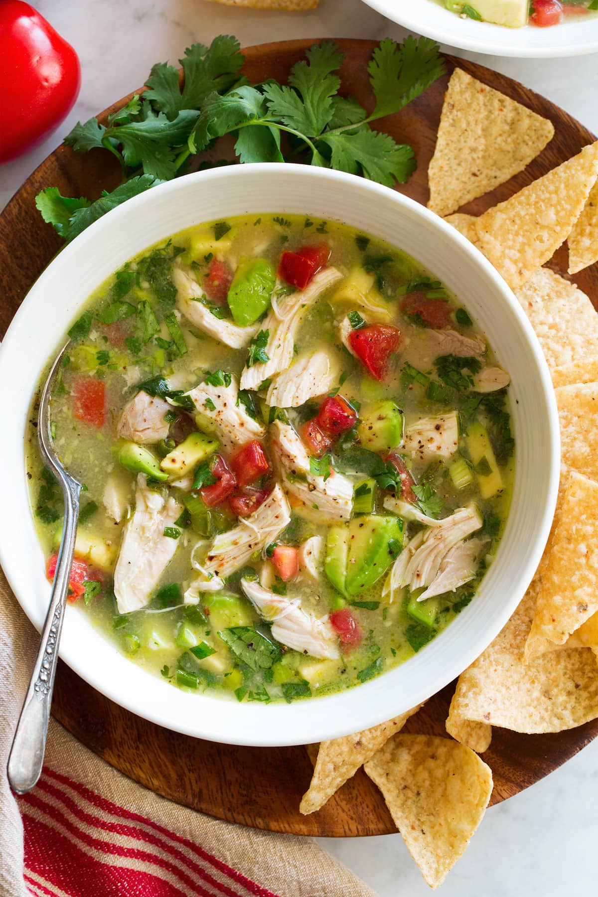 Chicken and avocado soup in a serving bowl set over a wooden plate. Shown overhead with a side of tortilla chips and cilantro.