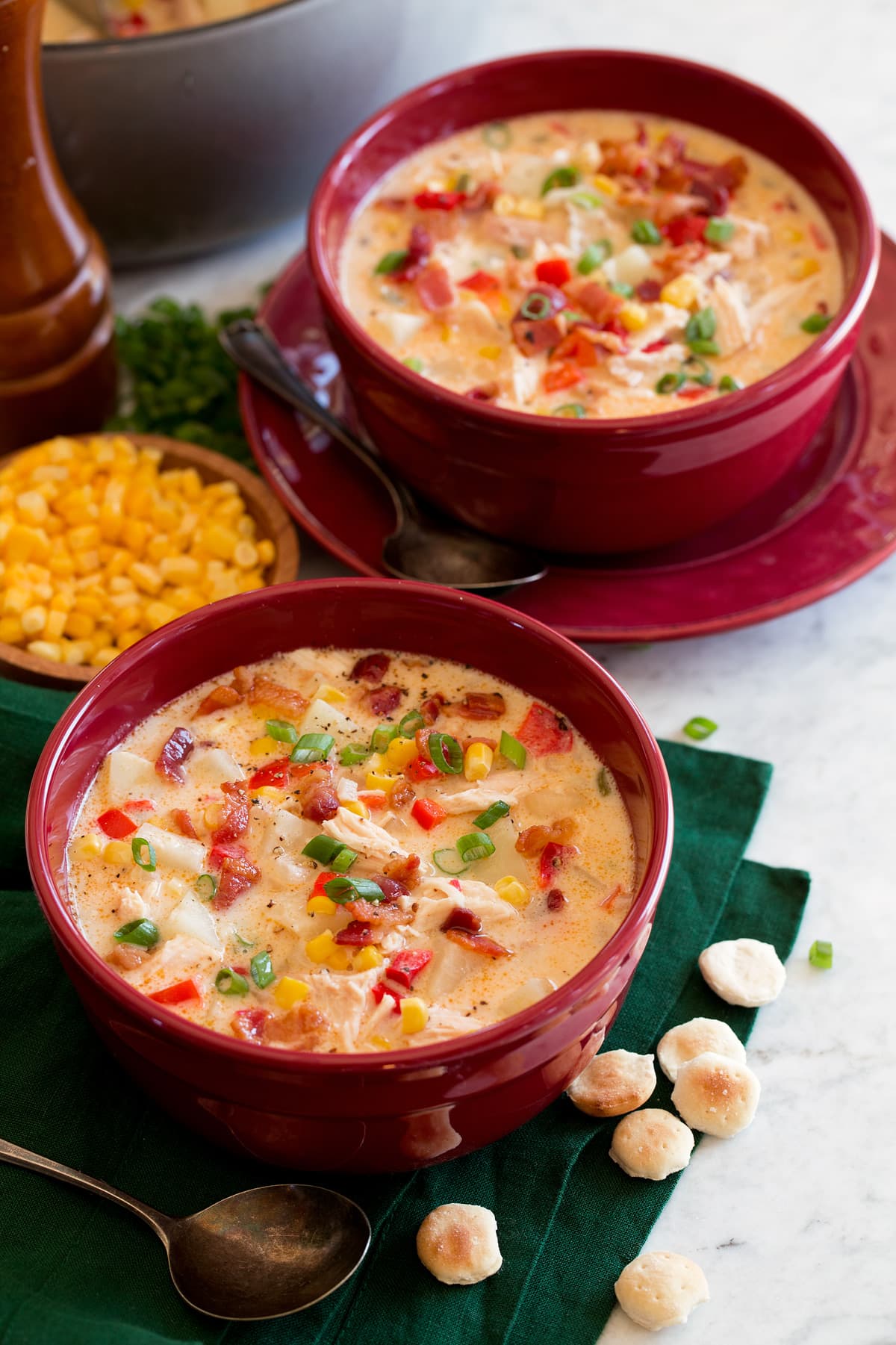 Two servings of chicken corn chowder in red bowls sitting over a green cloth on a marble surface.