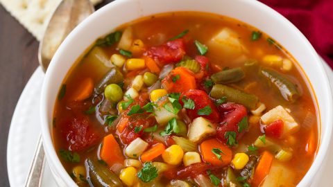 Single serving of vegetable soup in a white bowl set over a white plate. Set on a dark wooden table with a red napkin to the side.