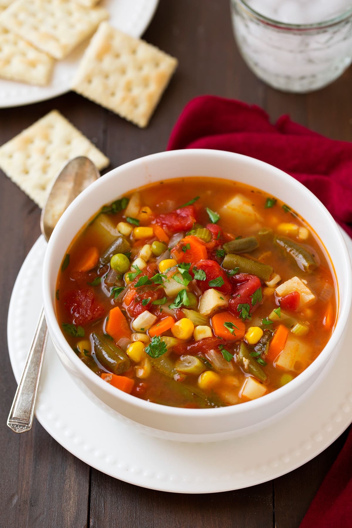 Single serving of healthy vegetable soup in a white bowl set over a white plate. Set on a dark wooden table with a red napkin to the side.