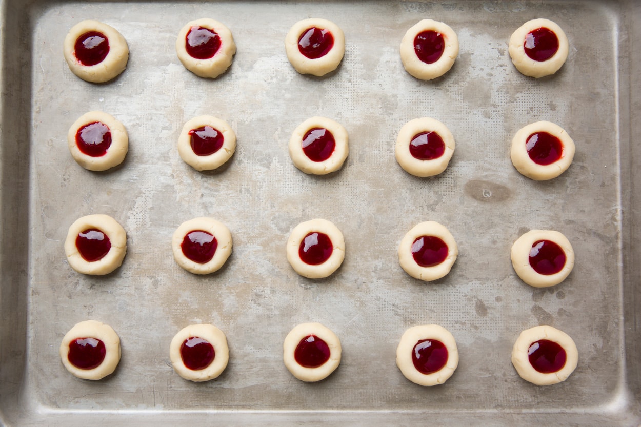 Showing how to make thumbprint cookies. Filling indented cookie dough on baking sheet with raspberry jam.