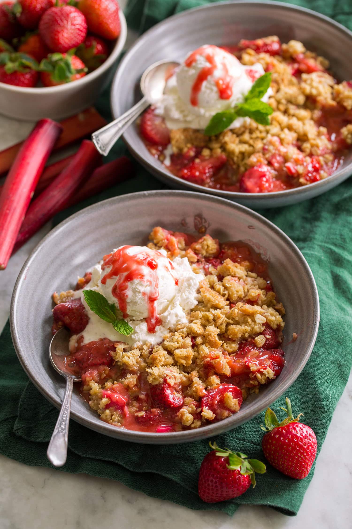 Two servings of strawberry rhubarb crisp with ice cream shown in grey bowls over green cloths.