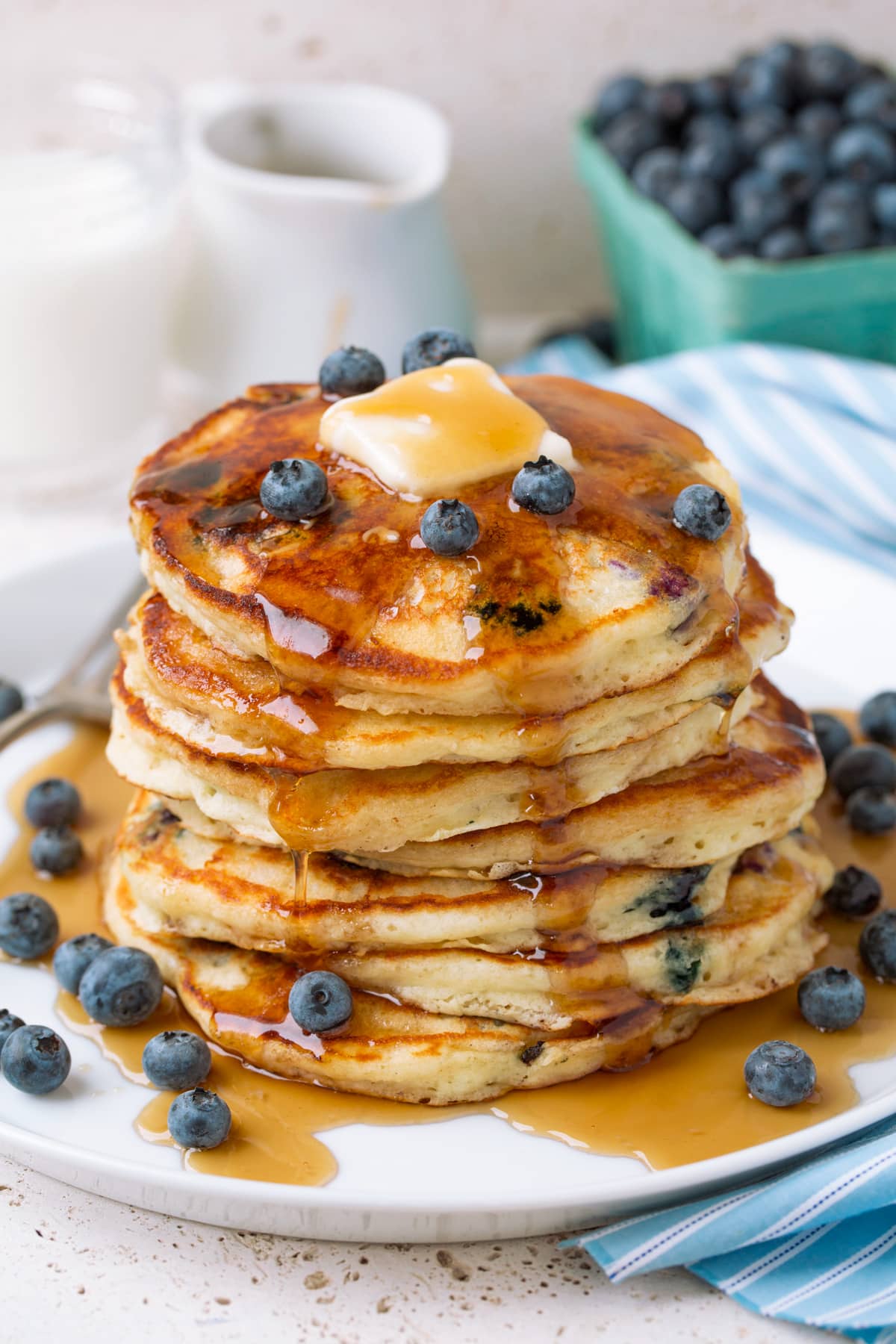 Stack of blueberry pancakes topped with blueberries, butter and maple syrup on a white serving plate.