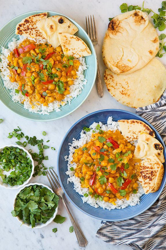 Two servings of vegetable curry one in a turquoise bowl and blue bowl. Each is served over basmati rice with a side of naan and garnished with cilantro.