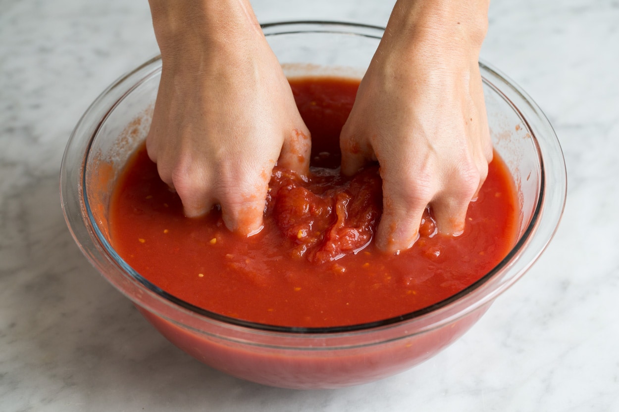 Crushing tomatoes by hand in a glass mixing bowl. Showing how to make marinara sauce.