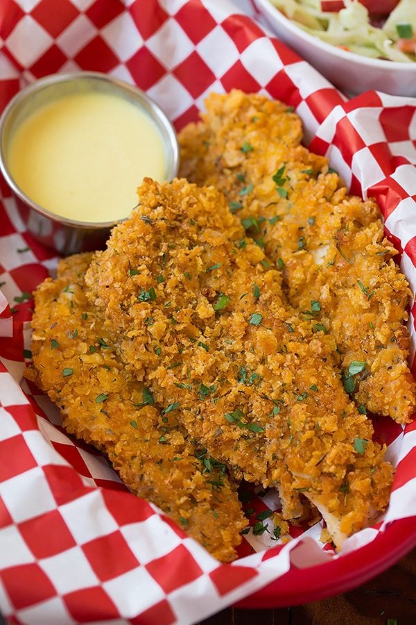 Three chicken strips laying in red plastic basket with a red checkered parchment paper liner. Honey mustard sauce is resting in a cup on the side.