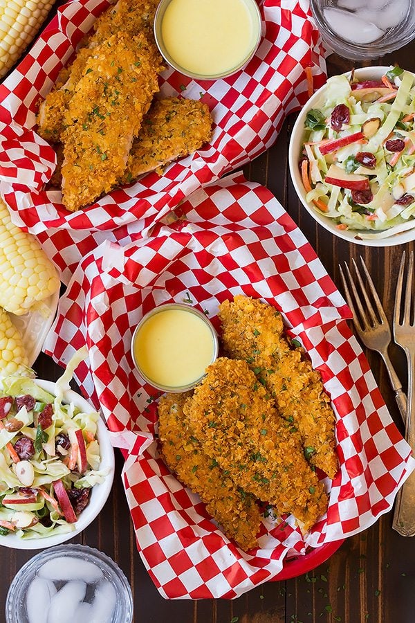 Two servings of baked chicken strips shown here along with a side of coleslaw and corn on the cob.