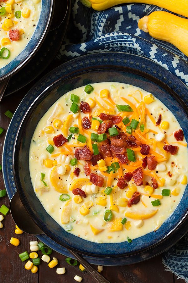 Summer Squash and Corn Chowder in a blue serving bowl set over a blue plate. It's garnished with shredded cheddar, bacon and green onions.