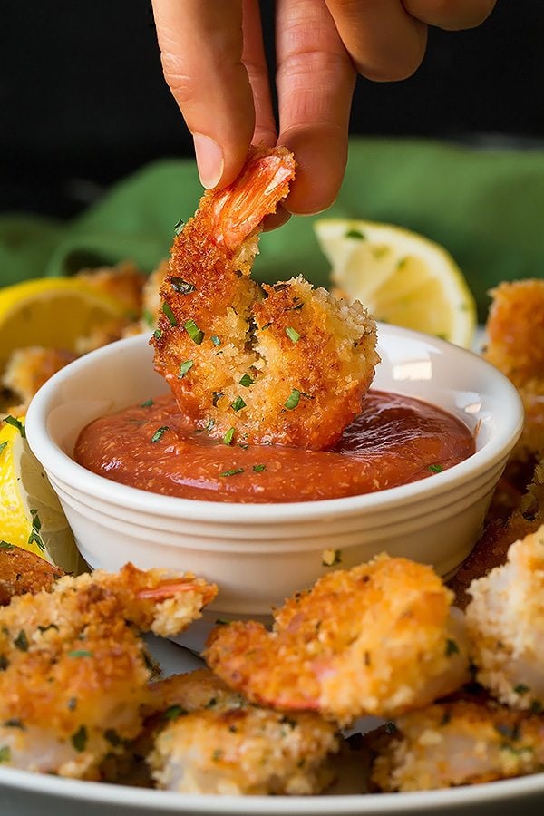 Cooked shrimp coated with panko being dipped in cocktail sauce in a white small bowl.