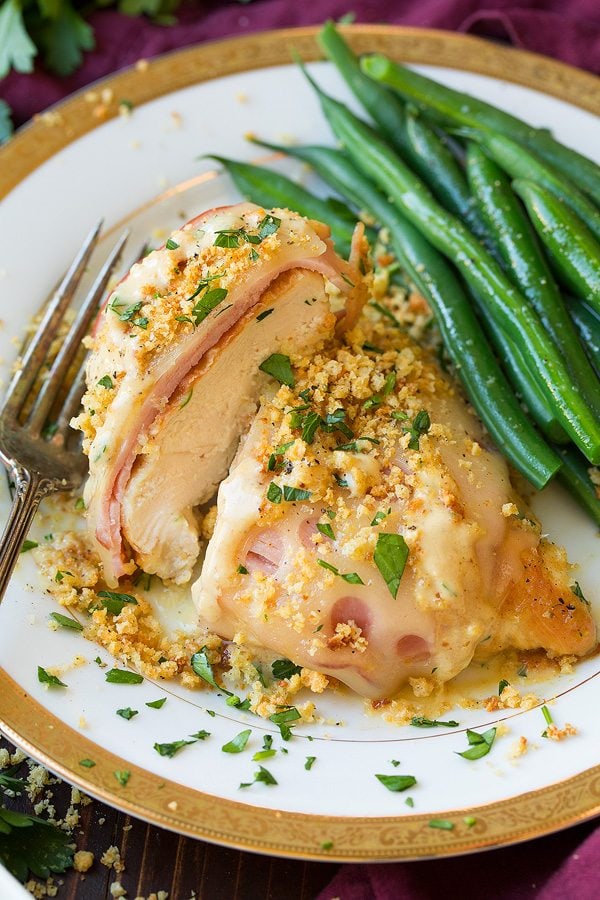 Chicken Cordon Bleu layered with thin deli ham slices, swiss cheese, croutons and parmesan. Served on a white plate with a gold rim with a side of steamed green beans.