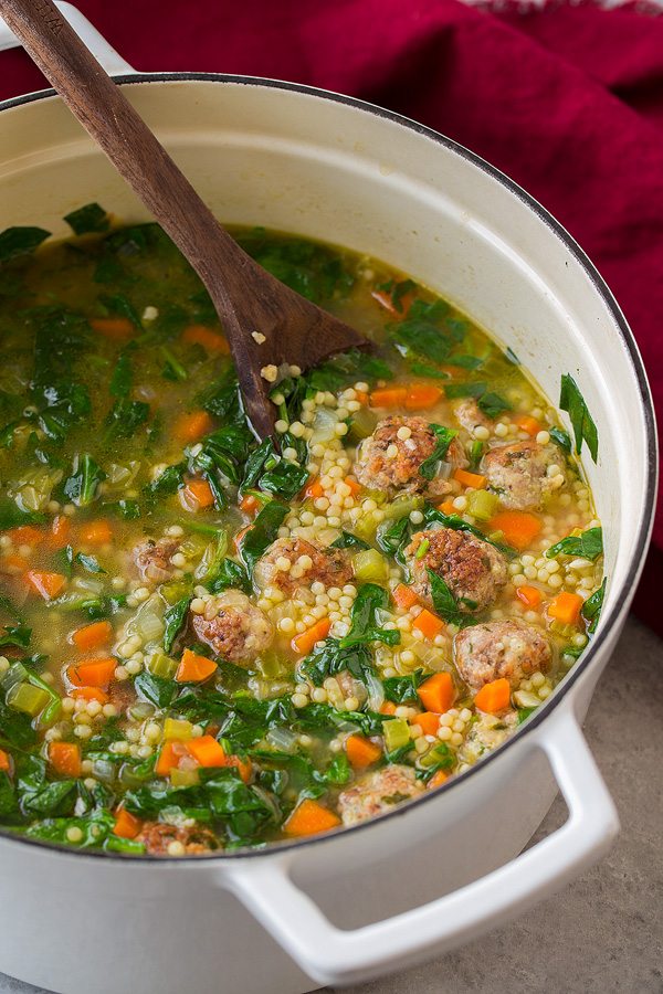 Italian Wedding Soup in a large white enameled pot. Soup includes meatballs, bits of pasta, carrots, celery, spinach and broth. 