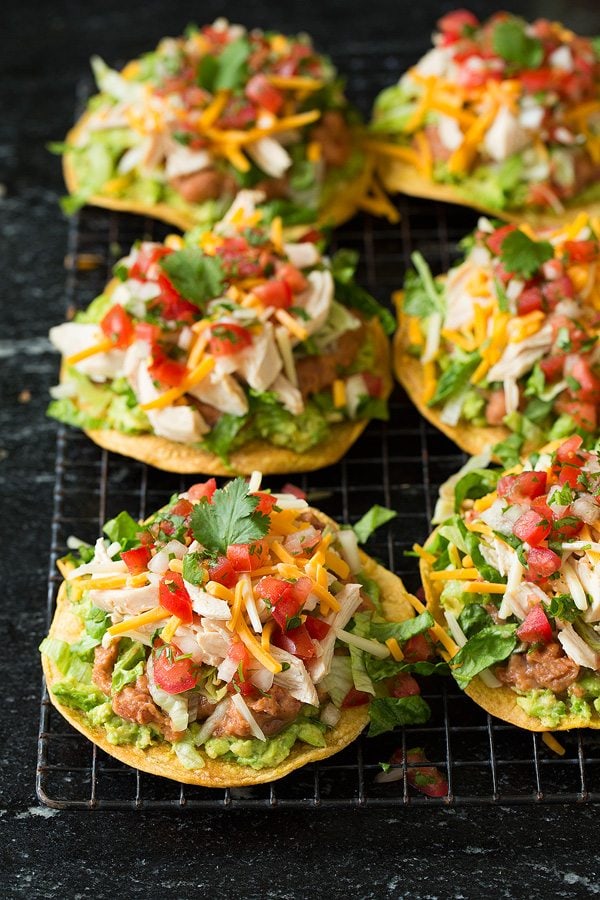 Six chicken tostadas are sitting on a wire cooling rack set over a black granite surface. Tostadas are layered with guacamole, refried beans, shredded cheese, pico de gallo and cilantro.
