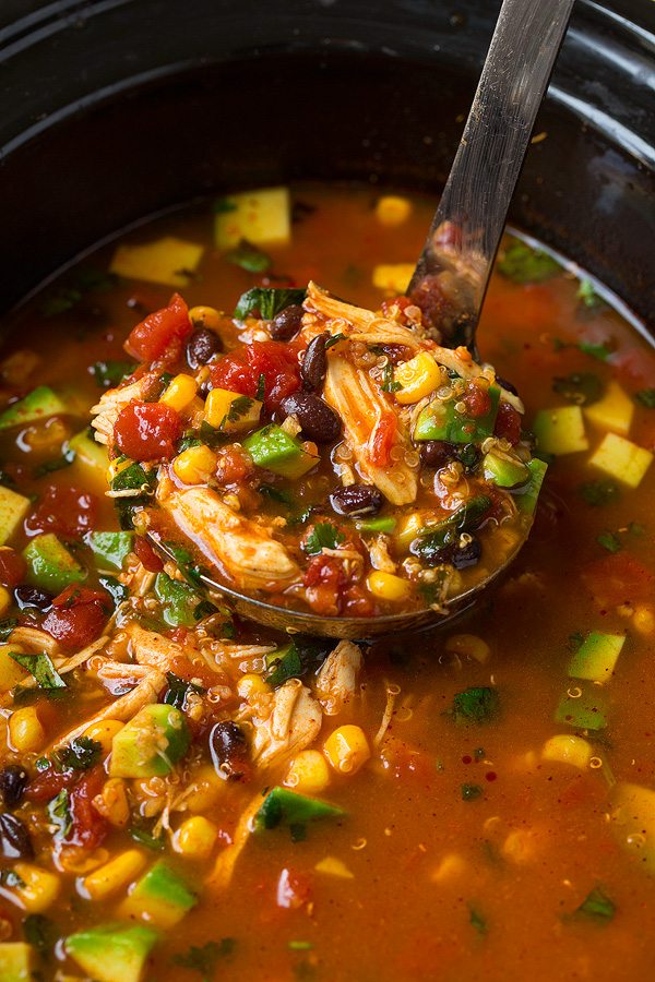 Close up image of chicken enchilada soup in a ladle in center of slow cooker. Ingredients showing include shredded chicken, black beans, corn, tomatoes, avocado and spiced broth.