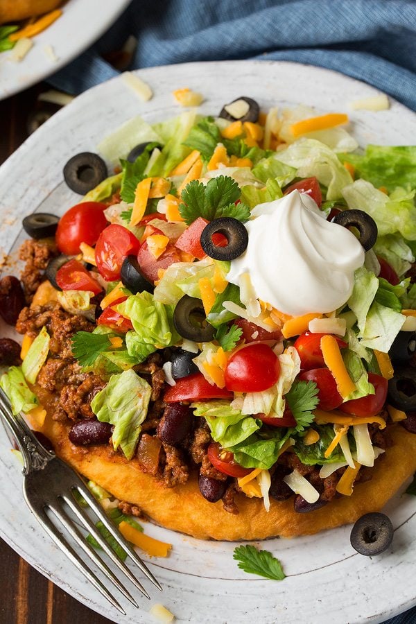 A homemade fry bread is layered with Navajo taco filling including seasoned ground beef, kidney beans, tomatoes, lettuce, olives, cheese and sour cream. It's set on a single serve white plate over a brown wooden surface.