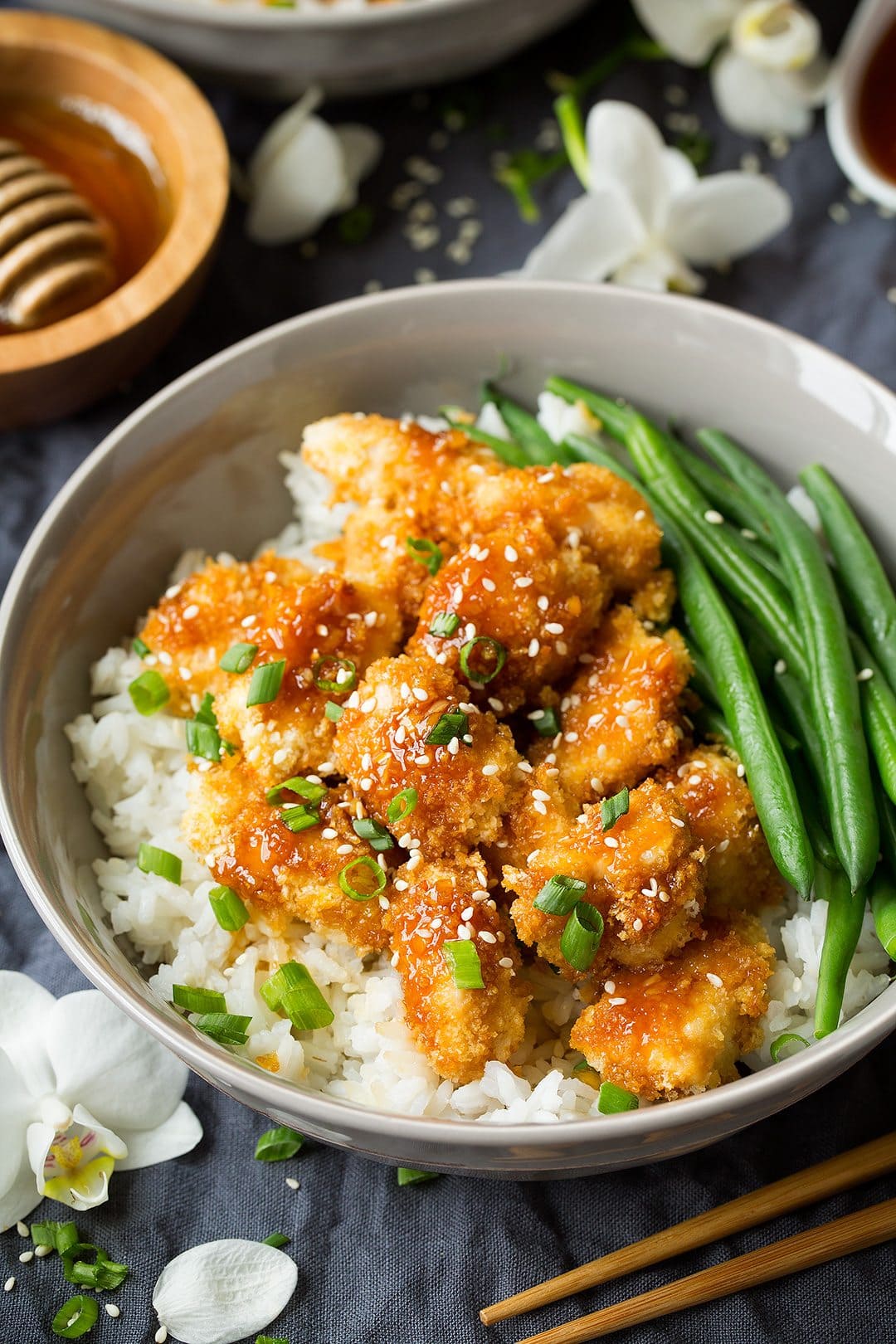 Honey Garlic Chicken set over a layer of rice in a grey bowl with a side of green beans. Bowl sitting on a grey tablecloth and decorated with white orchids.