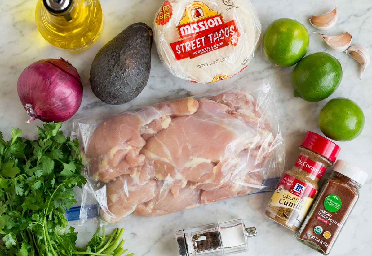 Image of ingredients used to make street tacos. Shows chicken thighs, cilantro, limes, garlic, avocado, chili powders, red onion, oil and tortillas. 