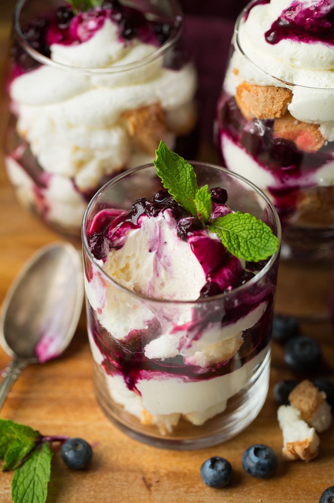 Image shows three trifles. Closes trifle is showing a scoop missing from it to show layers including angel food cake, cheesecake topping and blueberry sauce.