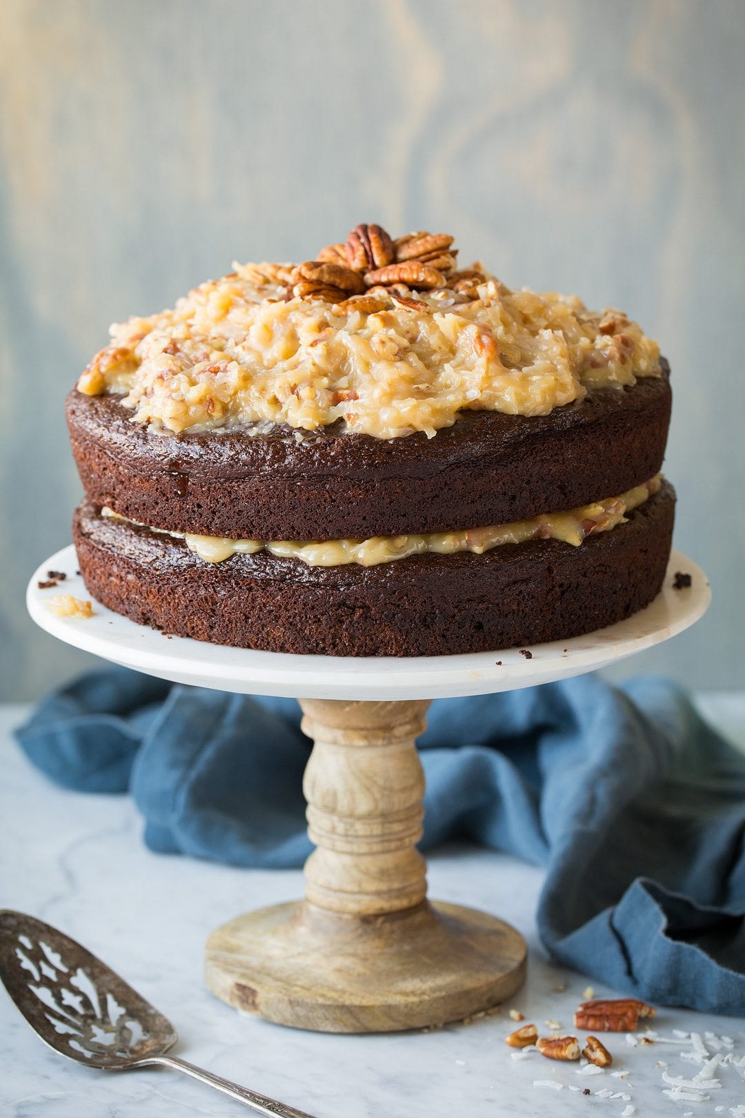 Whole two layer German Chocolate Cake on a marble cake stand with a light blue wooden background. 