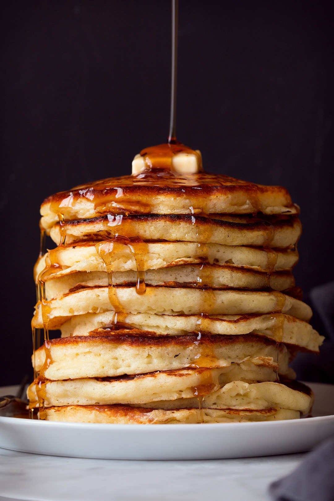 Maple syrup being poured over a tall stack of pancakes with a dab of butter on top. Pancakes are sitting on a large white plate on a marble surface, background is black.