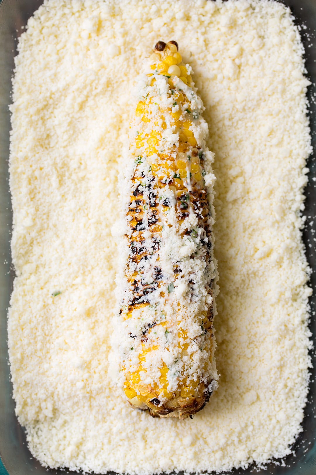 Corn cob being rolled in cotija in a shallow dish to make Mexican street corn.