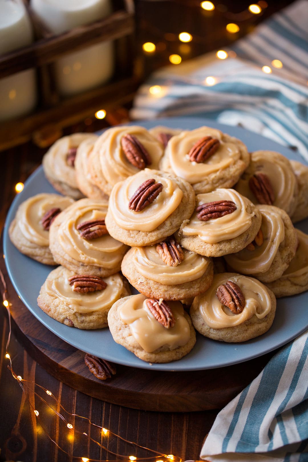 A pile of pecan cookies on a blue plate with christmas lights shown around plate and milk in the background.