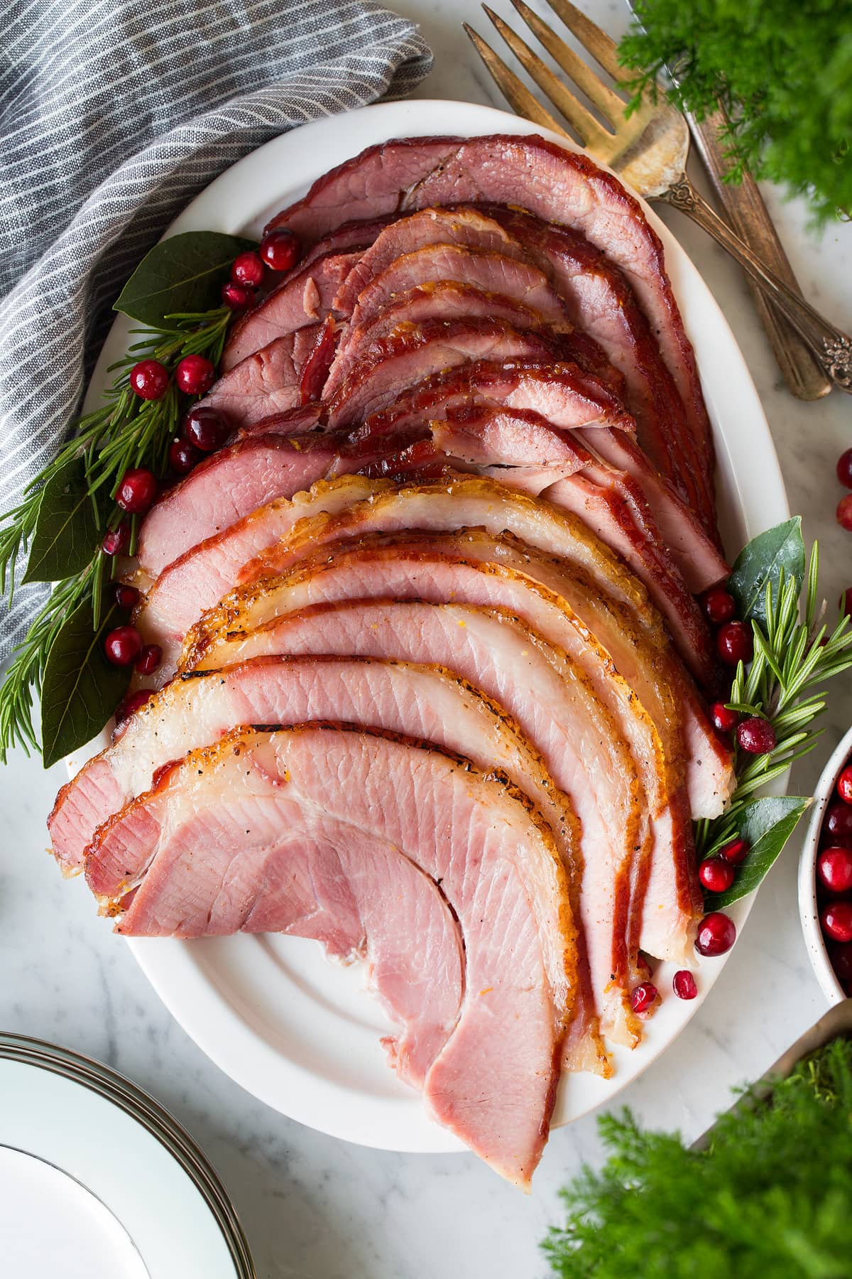 Honey baked ham slices on white serving platter garnished with fresh herbs and cranberries
