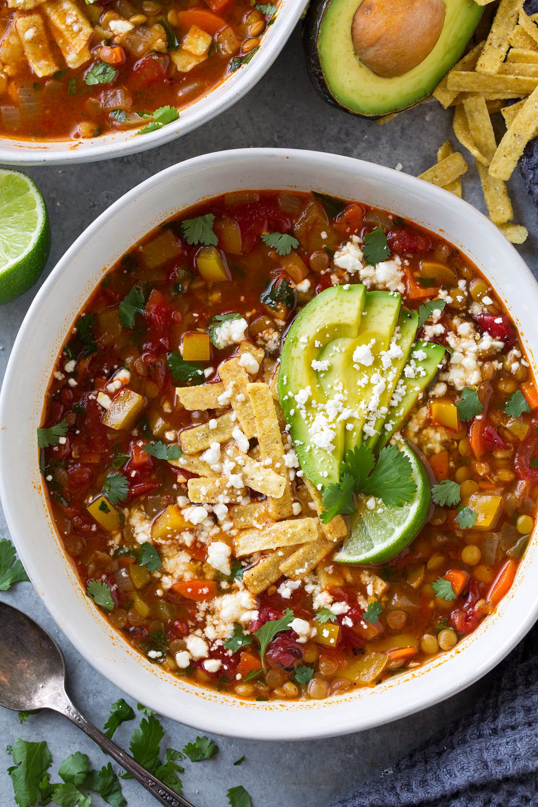 Mexican Lentil Soup in a white single serving bowl shown here up close to show the vegetables and variety of the texture in the soup. It's garnished with tortilla strips avocado slices and cheese