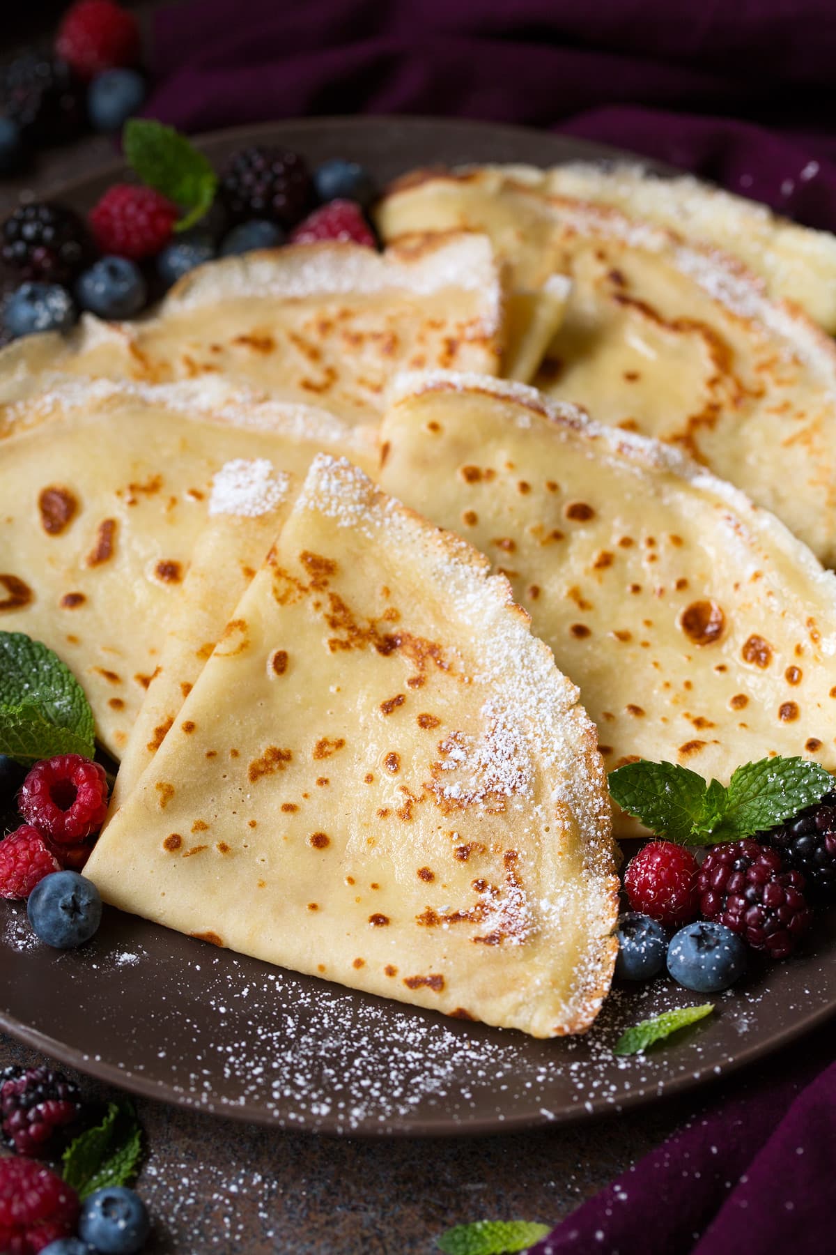 Best Crepes Recipe. Showing row of 6 crepes wrapped into quarters and dusted with powdered sugar, crepes are set on a dark brown serving plate, they're dusted with powdered sugar and served with a side of berries and mint.
