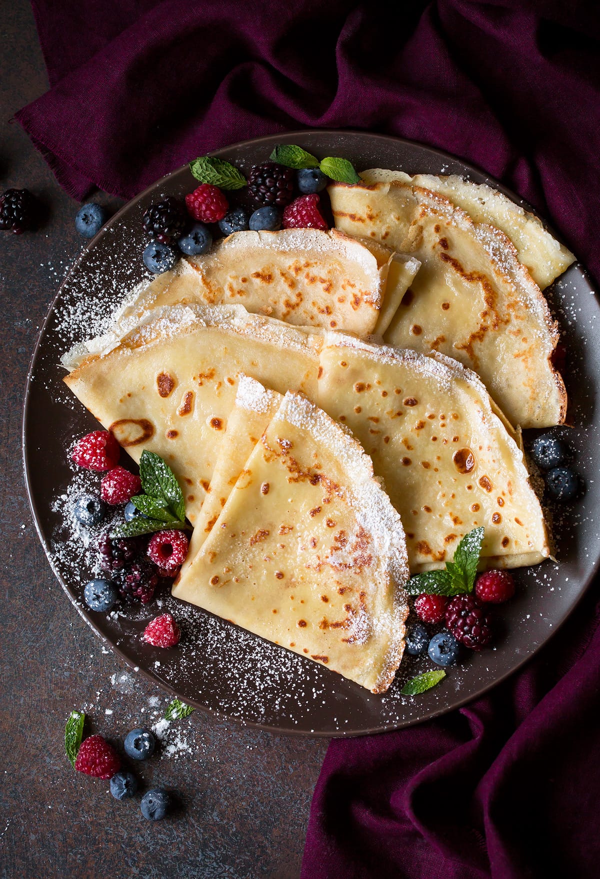Overhead image of plate full of folded crepes on a dark plate set over a maroon napkin.