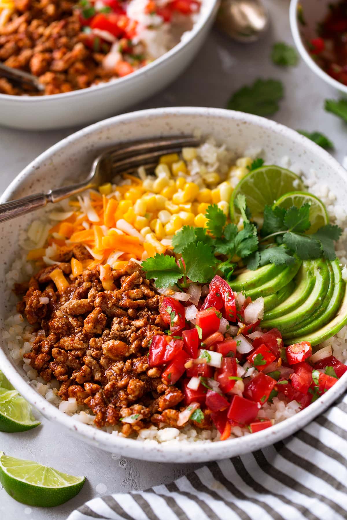 Taco Bowls shown in single serving layered with cauliflower rice, seasoned ground turkey, pico de gallo, shredded cheese, corn, avocado, cilantro and lime