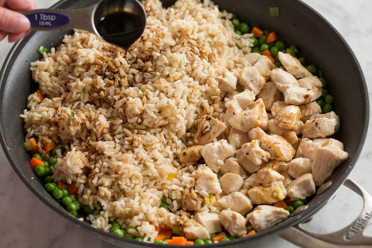 Pouring soy sauce over brown rice and chicken pieces in a large skillet to finish fried rice.