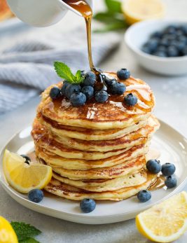 stack of eight lemon ricotta pancakes topped with fresh blueberries and mint maple syrup poured over pancakes lemon slices added on plate for garnish