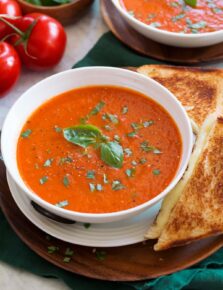 Serving of tomato basil soup shown close up in a white bowl with a grilled cheese to the side.