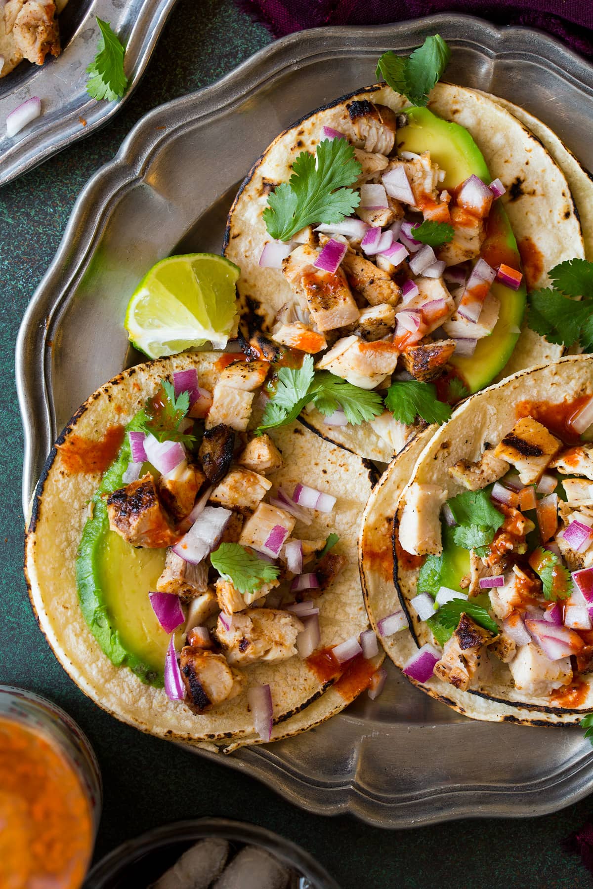 Image of 3 chicken street tacos on a metal plate. Tacos are made up of mini corn tortillas, grilled chicken, onion, cilantro, lime, avocado and Mexican hot sauce.