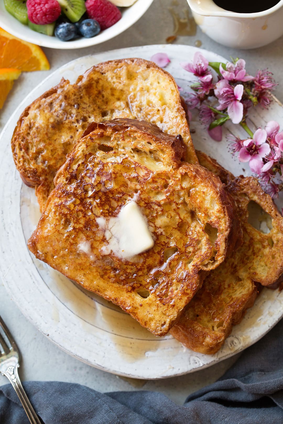 Three slices of french toast laying together on a white plate. Topped with butter and maple syrup and garnished with flowers on the side and fresh fruit.